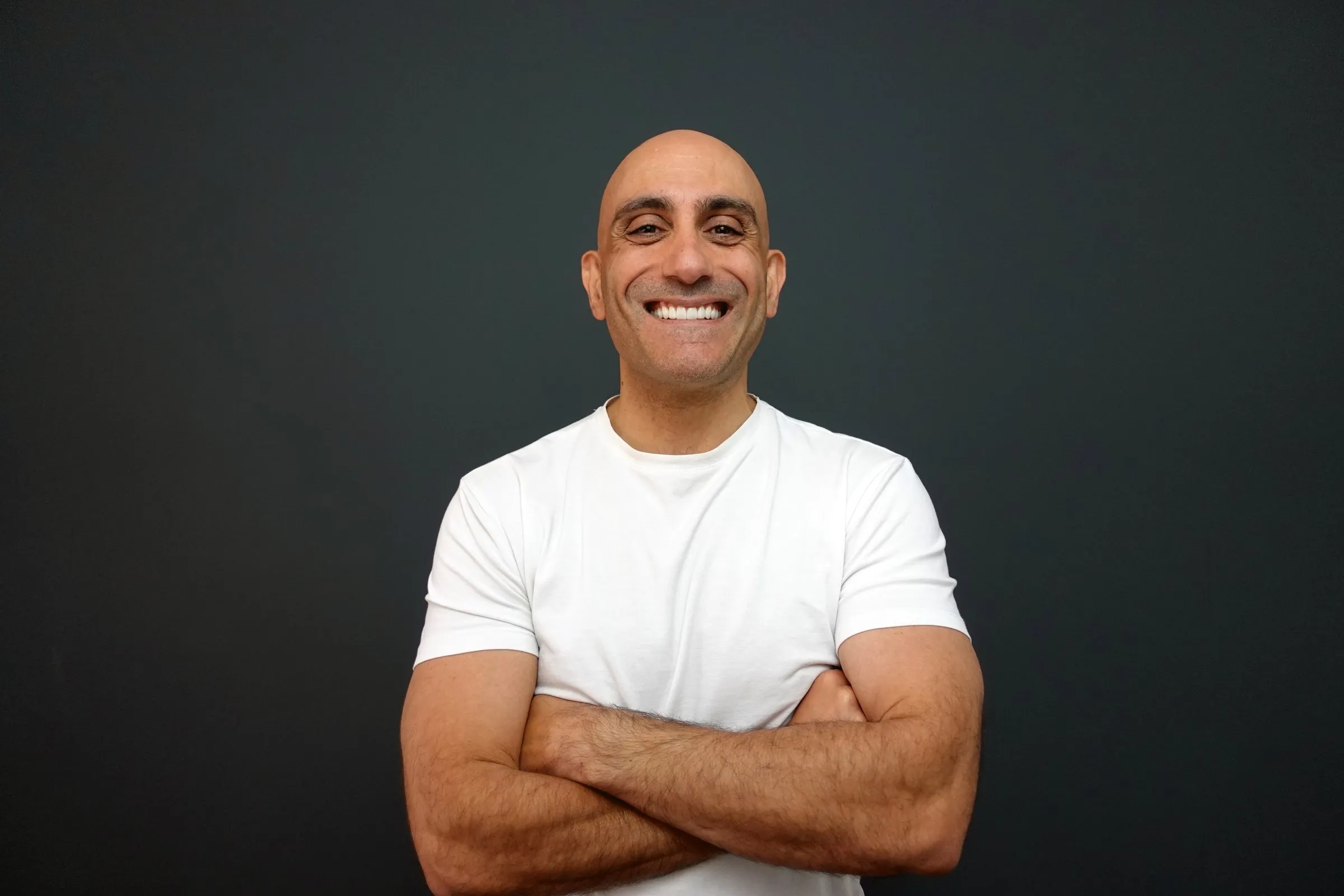 Chris K in a white T-shirt, arms crossed and smiling, against a dark studio backdrop