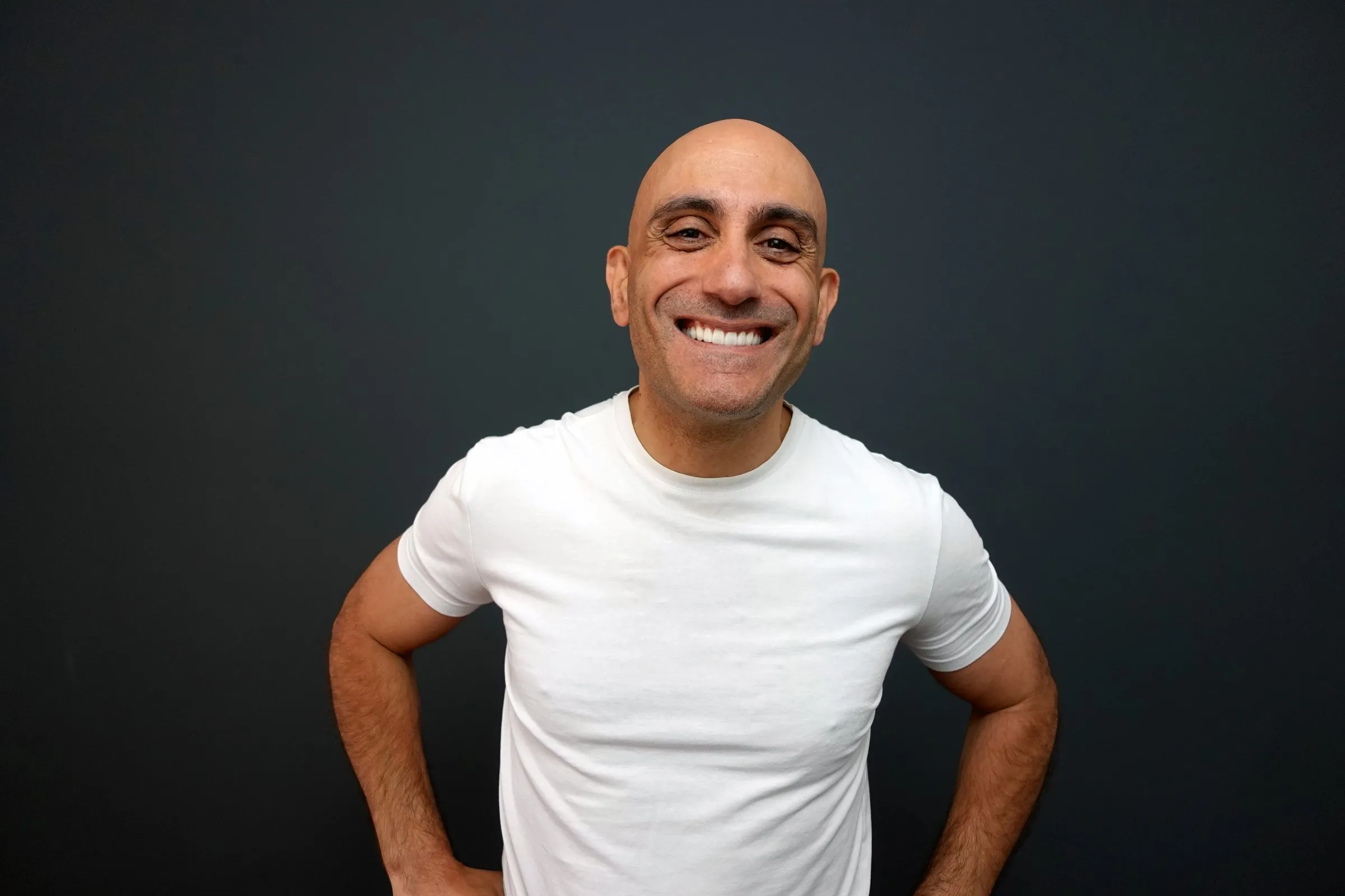 Chris K smiling, hands on hips, in a white T-shirt against a dark studio backdrop